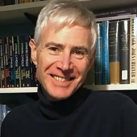 A photograph of Simon Mason, a white man smiling with white hair in front of a book shelf