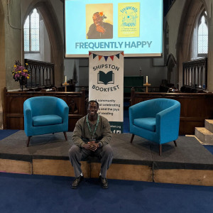 David Larbi sits on a stage with two blue chairs behind him
