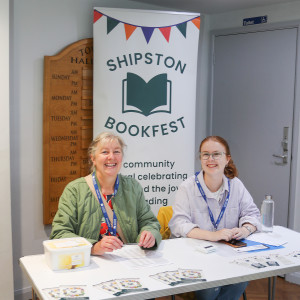 Two Bookfest volunteers sit at a table, smiling at the camera in front a colourful Bookfest banner.